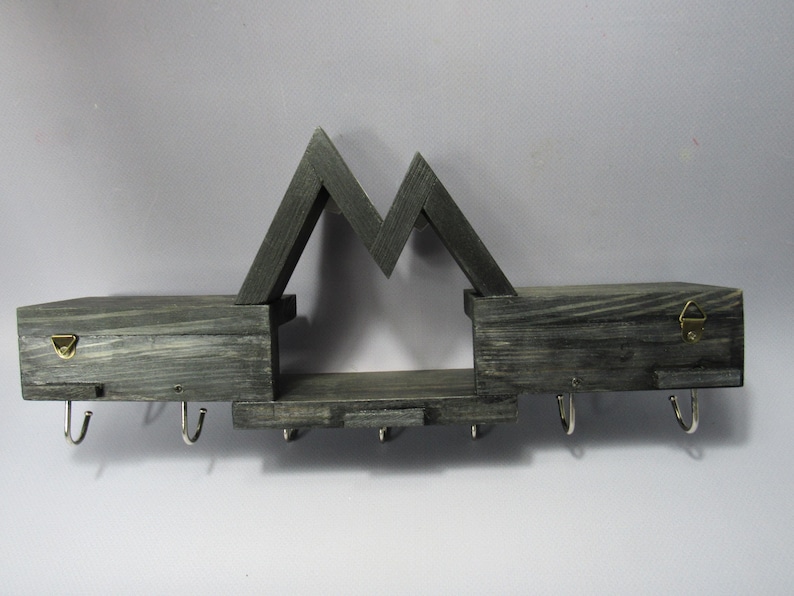 May include: A dark gray stained wooden wall shelf featuring a mountain-shaped silhouette. The unit includes two flat storage surfaces, a central lower shelf, and seven silver-toned metal hooks attached to the bottom for hanging items.