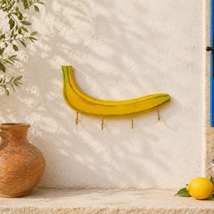 Può includere: Un eccentrico gancio da parete a forma di banana, dipinto di giallo con dettagli verdi, con quattro ganci in ottone. L'oggetto decorativo è montato su un muro bianco strutturato, con un vaso rustico e limoni nelle vicinanze.