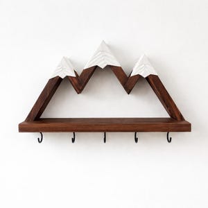 May include: A wall-mounted shelf shaped like a mountain range with three peaks. The structure is made of dark brown wood with white painted tips on the peaks. A flat horizontal shelf extends across the base, featuring five black metal hooks.