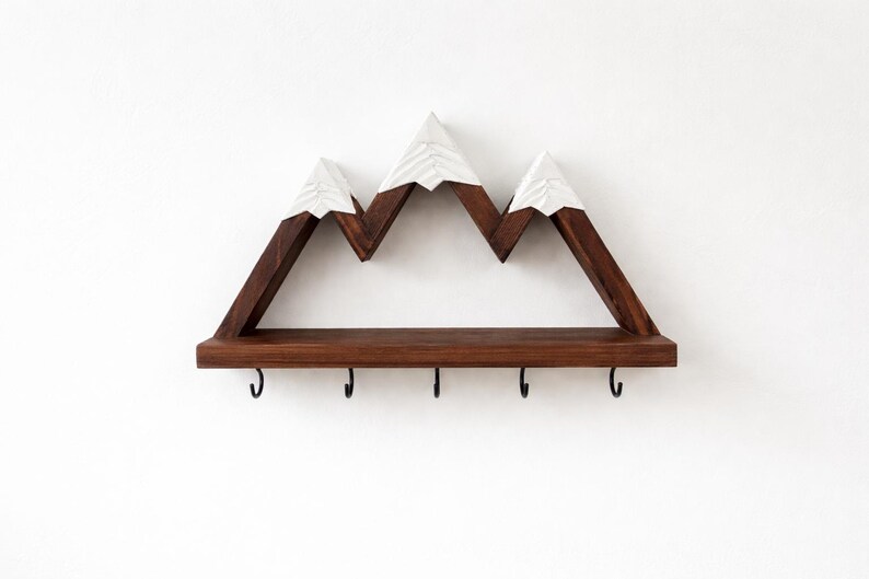 May include: A wall-mounted shelf shaped like a mountain range with three peaks. The structure is made of dark brown wood with white painted tips on the peaks. A flat horizontal shelf extends across the base, featuring five black metal hooks.