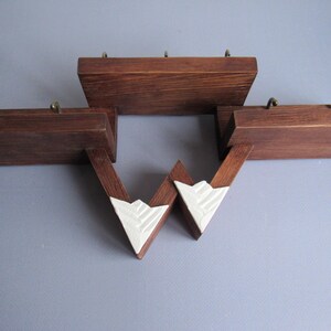 May include: A wall-mounted wooden shelf unit featuring three distinct horizontal platforms in a dark brown finish. Each platform is equipped with small brass-colored metal hooks. Two white, triangular, textured accents connect the lower sections.