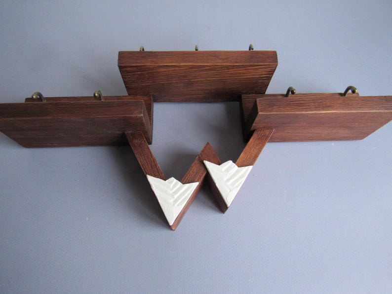 May include: A wall-mounted wooden shelf unit featuring three distinct horizontal platforms in a dark brown finish. Each platform is equipped with small brass-colored metal hooks. Two white, triangular, textured accents connect the lower sections.