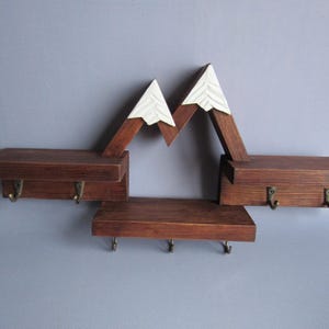 May include: A wall-mounted wooden shelf unit featuring a mountain-shaped design with white-painted peaks. The dark brown stained wood structure includes three horizontal shelves and seven small metal hooks for hanging items underneath the shelves.