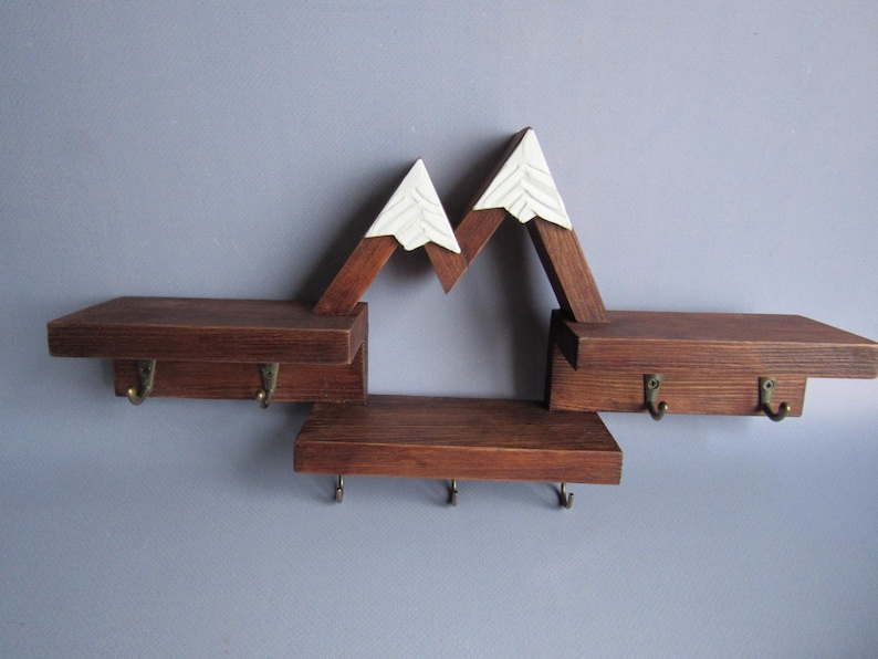 May include: A wall-mounted wooden shelf unit featuring a mountain-shaped design with white-painted peaks. The dark brown stained wood structure includes three horizontal shelves and seven small metal hooks for hanging items underneath the shelves.