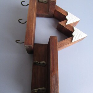May include: A wall-mounted wooden key rack featuring a dark brown stained finish and a mountain-shaped design. The structure includes two white-painted triangular peaks and five small, dark metal hooks attached to the vertical wooden panels.