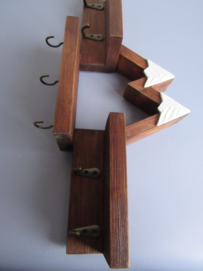 May include: A wall-mounted wooden key rack featuring a dark brown stained finish and a mountain-shaped design. The structure includes two white-painted triangular peaks and five small, dark metal hooks attached to the vertical wooden panels.