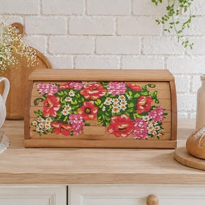 May include: Wooden bread box with a roll-top door, adorned with a vibrant floral design in shades of red, pink, and white. The bread box sits on a light-colored wooden countertop, with a loaf of bread and a white ceramic jar in the background.