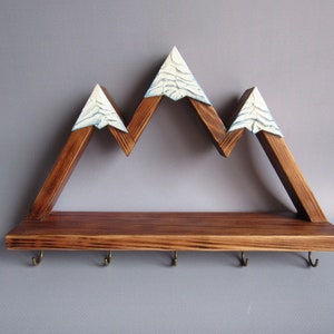 May include: Wooden mountain-shaped shelf with a brown stain and white snow-capped peaks. The shelf has a flat top with five hooks underneath.