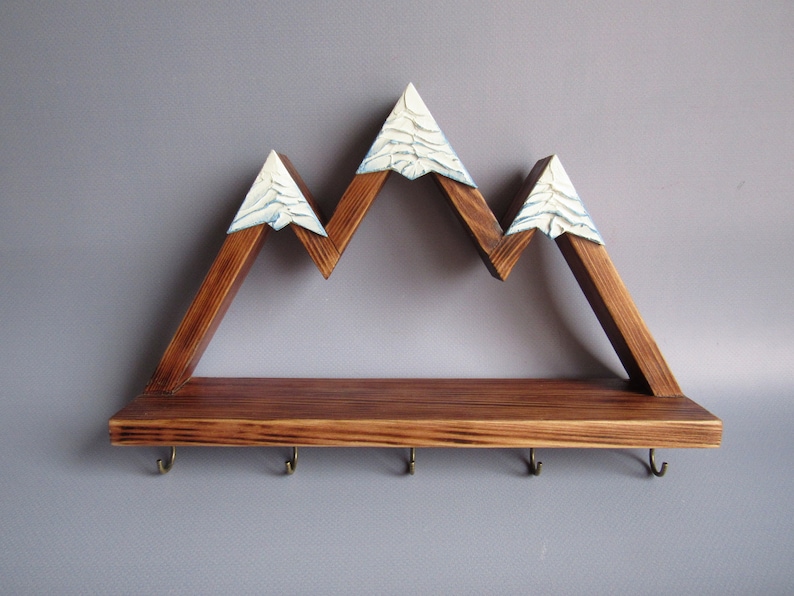 May include: Wooden mountain-shaped shelf with a brown stain and white snow-capped peaks. The shelf has a flat top with five hooks underneath.