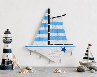 Könnte beinhalten: Ein hölzerner Wand-Schlüsselhalter in Form eines Segelboots mit weißen und blauen horizontalen Streifen auf dem Segel, einem schwarzen Mast und einem weißen Rumpf mit einem blauen Seestern-Akzent. Das Segelboot hat vier Metallhaken.