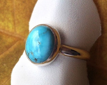 Op de afbeelding: Een zilveren ring met een ovale turquoise edelsteen gezet in een bezel setting. De ring wordt getoond op een witte ringhouder.