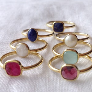 Könnte beinhalten: Ein Satz von sieben goldenen Ringen mit verschiedenen farbigen Edelsteinen. Die Ringe haben eine Vielzahl von Formen und Größen von Edelsteinen, darunter runde, ovale und quadratische. Die Edelsteine sind in Goldfassungen gefasst und von einem dünnen Goldband umgeben.