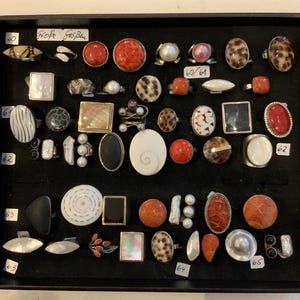 Pode incluir: Uma coleção de anéis variados exibidos numa bandeja preta, com diversas formas e materiais, incluindo coral vermelho, madrepérola branca, ónix preto e cabochões de concha manchada em aros de metal, com pequenas etiquetas numeradas de 60 a 65.