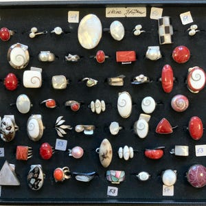May include: A black display tray containing a collection of assorted rings featuring various gemstones, shell inlays, and metal settings. The rings are arranged in rows, with small white paper labels marked with numbers placed next to several items.