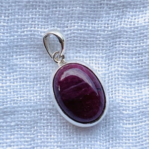Può includere: Un ciondolo d'argento con una pietra ovale di colore viola scuro. Il ciondolo ha un design semplice ed è perfetto per aggiungere un tocco di eleganza a qualsiasi outfit.