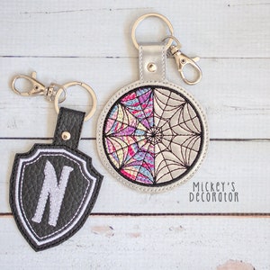 May include: Two embroidered keychains. One is a black shield with a white letter N. The other is a round, silver keychain with a colorful spiderweb design. The text "Mickey's decorator" is visible in the bottom right corner.