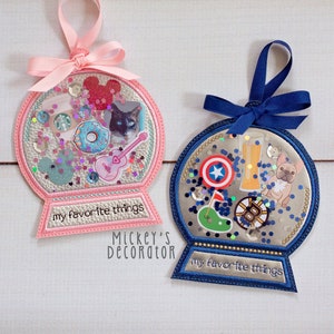 May include: Two pink and blue felt ornaments shaped like snow globes with glitter and various images inside. The ornaments have a white background with the text "my favorite things" on the bottom. The pink ornament has images of a coffee cup, a donut, a cat, a guitar, and a Mickey Mouse head. The blue ornament has images of a beer glass, a french bulldog, a golf club, a Captain America shield, and a Boston Bruins logo. The text "Mickey's Decorator" is printed below the ornaments.
