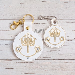 May include: Two white leather keychains with gold embroidery of a stylized tree with a crown on top. The keychains are on a white wooden background.  Mickey's Decoration is printed on the larger keychain.