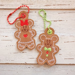 Może przedstawiać: Dwa skórzane breloczki w kształcie piernikowego ludzika z haftowanymi detalami. Jeden brelok jest brązowy z czerwoną kokardką i sercem, drugi jest brązowy z zieloną kokardką i zielonym guzikiem. Oba breloczki mają białe szwy i wiszą na metalowym łańcuszku.