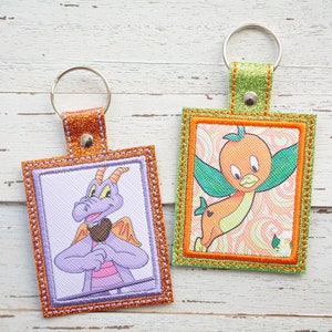 Puede incluir: Dos llaveros bordados con costuras naranja y verde. Un llavero presenta un dragón morado con una camisa rosa y una piruleta marrón. El otro llavero presenta un pájaro amarillo con alas naranjas y una hoja verde.