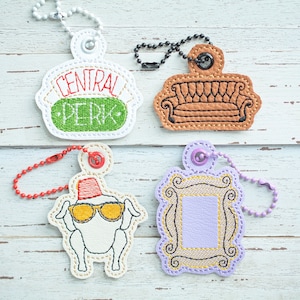 May include: Four embroidered keychains featuring iconic symbols from the TV show "Friends". The keychains are white with colorful embroidery. One keychain features the words "Central Perk", another features a purple frame, another features a brown couch, and the last features a turkey with a red fez.