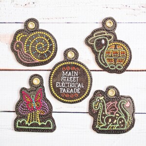 May include: Five embroidered keychains with a black background and colourful thread. The keychains feature a snail, a turtle, a butterfly, a dragon, and a circle with the text "Main Street Electrical Parade".