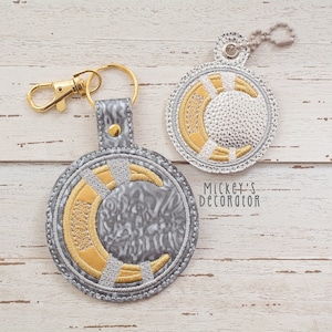 Op de afbeelding: Twee ronde sleutelhangers met een hemels ontwerp. Eén sleutelhanger heeft een goudkleurige sluiting, de andere een ketting. Beide hebben een grijs, goud en zilver geborduurd ontwerp met een halve maan en een zonnepatroon. De tekst "Mickey's Decorator" is zichtbaar.