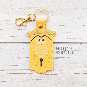 May include: Yellow leather keychain with an embroidered design of a character from the movie 'Beauty and the Beast'. The character has a mustache, large eyes, and a keyhole in its chest. The keychain also has a gold metal ring and a gold metal clasp. The text 'Mickey's Decorator' is visible in the bottom right corner of the image.