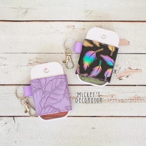 May include: Two hand sanitizer holders with keychains. One is lavender with a leaf pattern, the other is black with iridescent feather designs. Both have a white top and a purple ribbon loop.