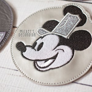 Puede incluir: Un parche redondo plateado con una imagen bordada en blanco y negro de Mickey Mouse con un sombrero de purpurina plateado. El texto "Mickey's Decorator" est&aacute; impreso en el parche.