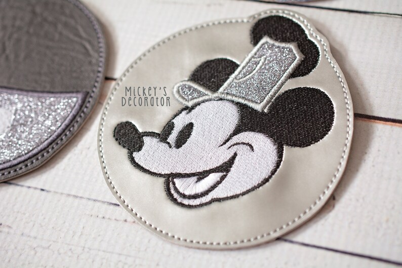 Puede incluir: Un parche redondo plateado con una imagen bordada en blanco y negro de Mickey Mouse con un sombrero de purpurina plateado. El texto "Mickey's Decorator" est&aacute; impreso en el parche.