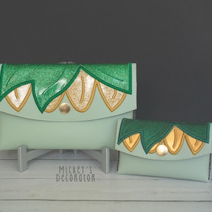 May include: Two green and gold clutches with a floral design. The larger clutch is on a stand and the smaller clutch is in front of it.