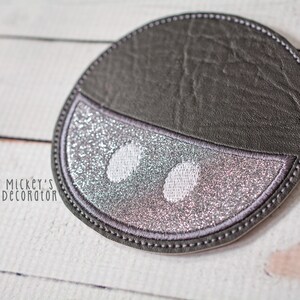 Puede incluir: Un posavasos redondo gris y plateado con purpurina con un dise&ntilde;o blanco cosido de dos ojos. El posavasos est&aacute; hecho de piel sint&eacute;tica y tiene un borde cosido.