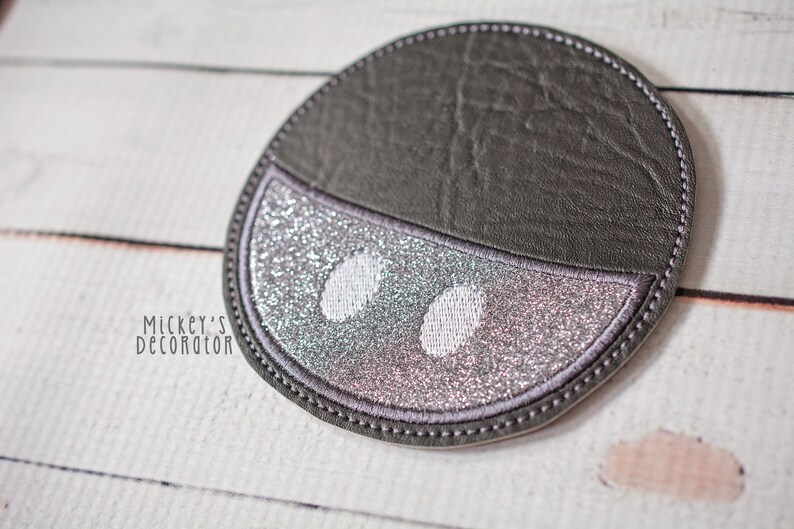 Puede incluir: Un posavasos redondo gris y plateado con purpurina con un dise&ntilde;o blanco cosido de dos ojos. El posavasos est&aacute; hecho de piel sint&eacute;tica y tiene un borde cosido.