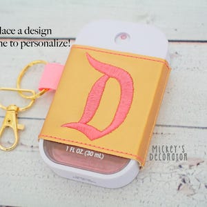 Puede incluir: Un estuche personalizado para botella de spray dorado con un dise&ntilde;o "D" rosa bordado. Incluye una correa rosa y un cierre dorado. La botella contiene 30 ml de l&iacute;quido. El texto en la imagen dice "&iexcl;Coloca un dise&ntilde;o o nombre para personalizar!"