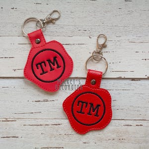 Könnte beinhalten: Zwei rote Schlüsselanhänger mit einem schwarzen kreisförmigen Design und den Buchstaben "TM" in der Mitte gestickt. Jeder Schlüsselanhänger hat einen silbernen Verschluss und einen Lederriemen. Die Schlüsselanhänger befinden sich auf einem hellen Holzgrund.