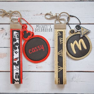 May include: Two keychains with black and white Mickey Mouse and Harry Potter themed fabric straps. The Mickey Mouse keychain has a red circle with the name "Cassy" embroidered in black. The Harry Potter keychain has a gold circle with the letter "M" embroidered in gold. Both keychains have silver metal rings.
