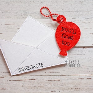May include: A white, stitched envelope with the text "SS GEORGIE" and a red balloon-shaped tag that reads "you'll float too". The tag is attached with a red chain. The items are on a light wood surface.
