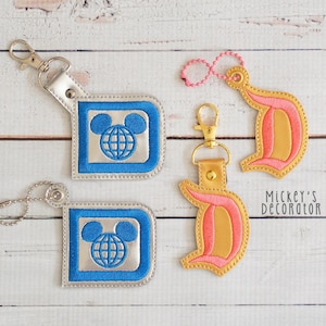 May include: Two keychains with embroidered designs. One keychain features a blue square with a Mickey Mouse head and a globe inside. The other keychain features a gold D with pink stitching. The text "Mickey's Decorator" is visible in the bottom right corner of the image.