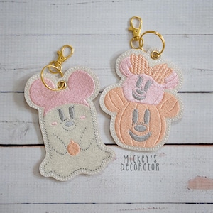 May include: Two embroidered keychains with gold-tone lobster clasps. One features a light grey ghost with pink ears holding a small orange pumpkin. The other shows a peach-coloured pumpkin with pink ears and a bow. Text reads Mickey's Decorator.