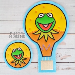 May include: Two felt appliques featuring a green frog in a hot air balloon. The larger applique is a hot air balloon with a yellow, orange, and purple design. The smaller applique is a circle with a yellow background and a green frog.