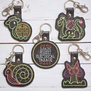 以下が含まれることがあります： 異なるデザインの刺繍されたキーホルダーが5つあります。キーホルダーは黒で、カラフルな刺繍が施されています。デザインには、カメ、ドラゴン、カタツムリ、蝶、そして「メインストリートエレクトリカルパレード」という文字が書かれた円形のものがあります。