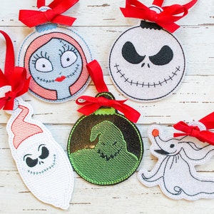 Op de afbeelding: Vijf kerstversieringen van vilt met personages uit de film Nightmare Before Christmas. De versieringen zijn wit met zwarte borduursels en bevatten Jack Skellington, Sally, Zero, Oogie Boogie en een kerstman-versie van Jack Skellington.