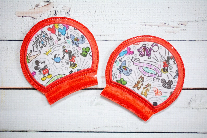 May include: Two red fabric Mickey Mouse ears with a white background featuring a variety of Disney characters and the text "Walt Disney World Disneyland".