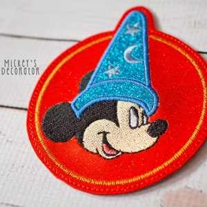 Puede incluir: Un parche bordado rojo con un contorno dorado que presenta a Mickey Mouse con un sombrero de mago azul con estrellas y una luna creciente. El parche tiene un di&aacute;metro de aproximadamente 7,5 cm.