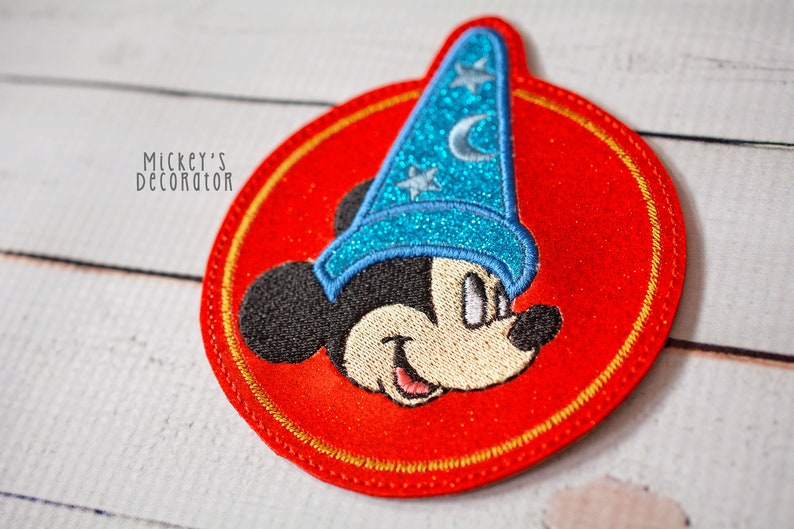Puede incluir: Un parche bordado rojo con un contorno dorado que presenta a Mickey Mouse con un sombrero de mago azul con estrellas y una luna creciente. El parche tiene un di&aacute;metro de aproximadamente 7,5 cm.