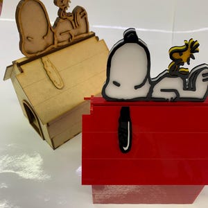 May include: Two Snoopy dog houses. One is a wooden dog house with Snoopy and Woodstock on top. The other is a red dog house with Snoopy and Woodstock on top. The Snoopy and Woodstock figures are black, white, and yellow.