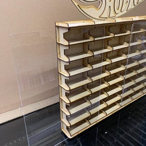 May include: A wooden display shelf with clear acrylic panels for storing Hot Wheels cars. The shelf has multiple rows of slots for displaying the cars. The shelf is made of natural wood and has a simple design.