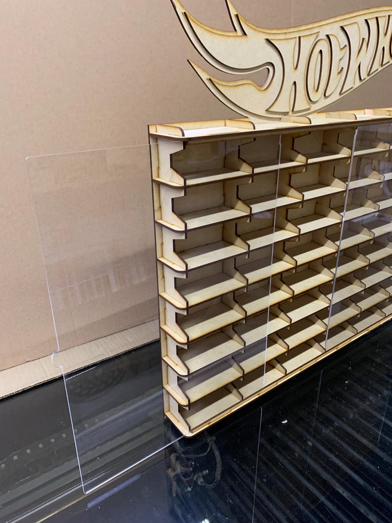 May include: A wooden display shelf with clear acrylic panels for storing Hot Wheels cars. The shelf has multiple rows of slots for displaying the cars. The shelf is made of natural wood and has a simple design.
