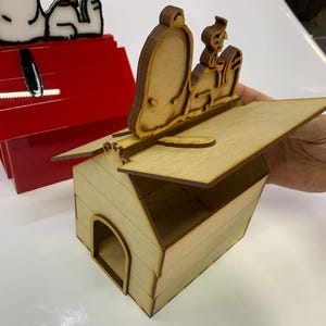May include: A wooden doghouse with a hinged roof, featuring a laser-cut Snoopy and Woodstock design. The doghouse is made of light-colored wood, with a contrasting red doghouse in the background. The design is a charming decorative item.